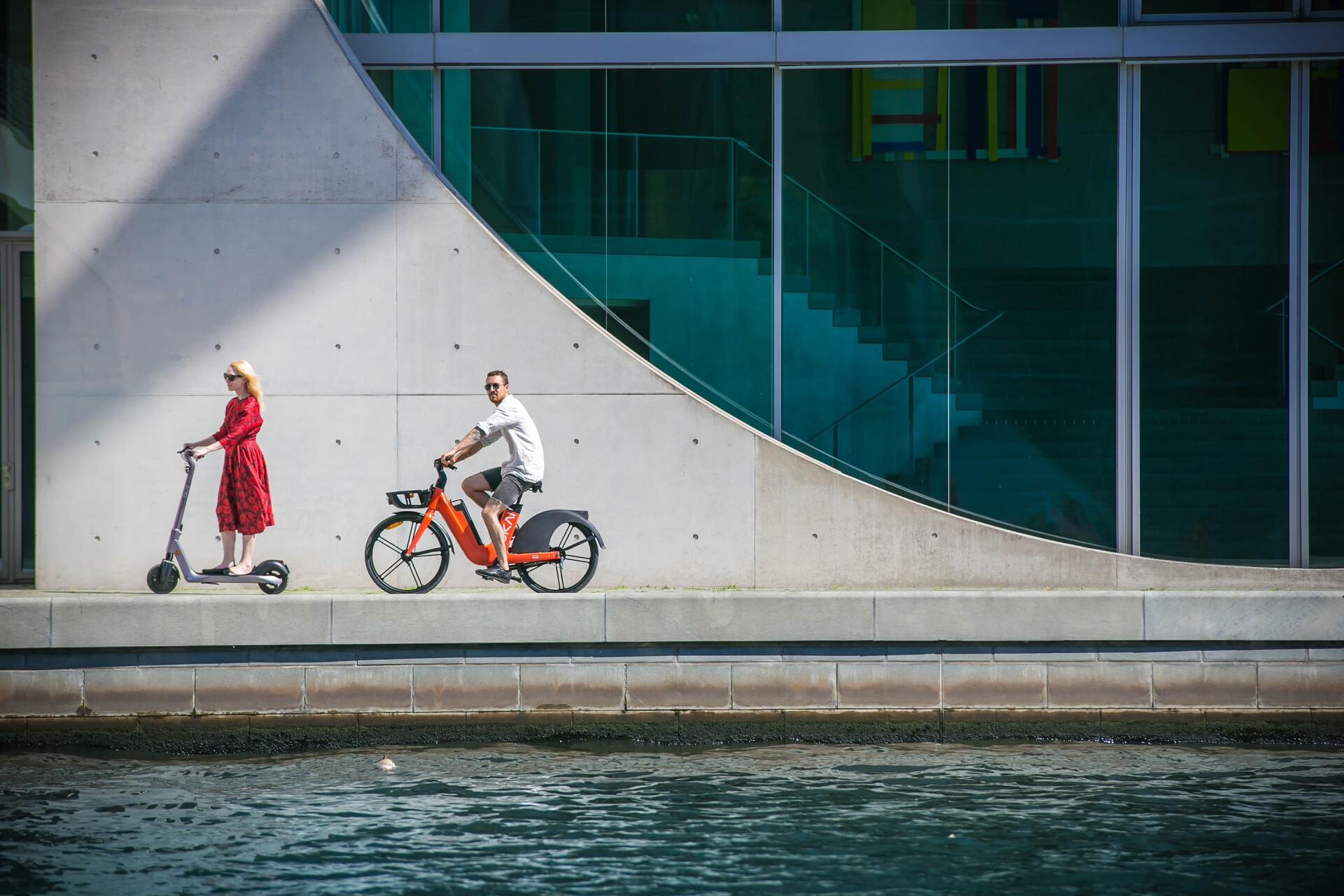 One caucasian female and one caucasian male riding a kick-scooter and an electric bike next to an architectural building.