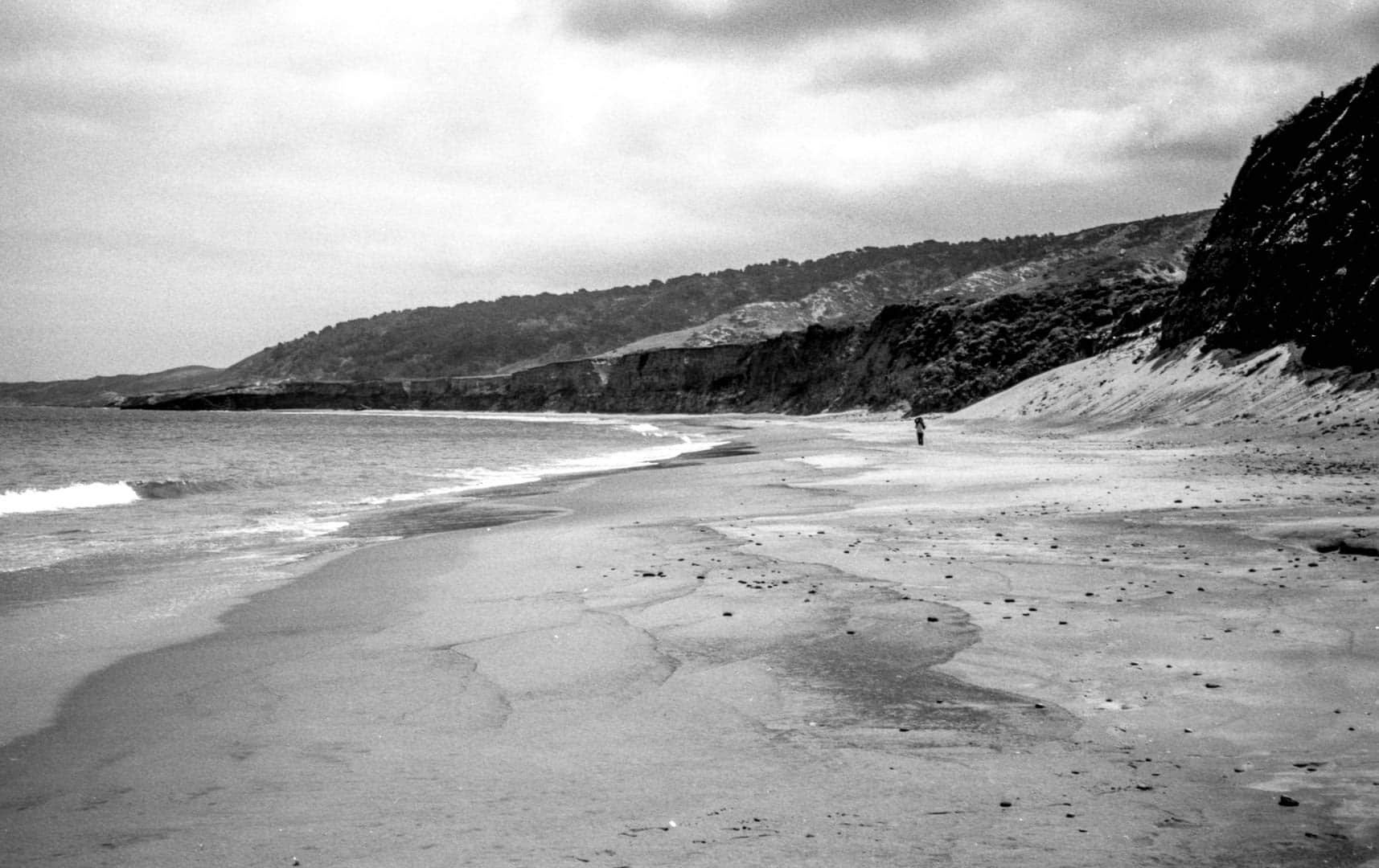 a windswept beach with a single person on it and steep dark bluffs