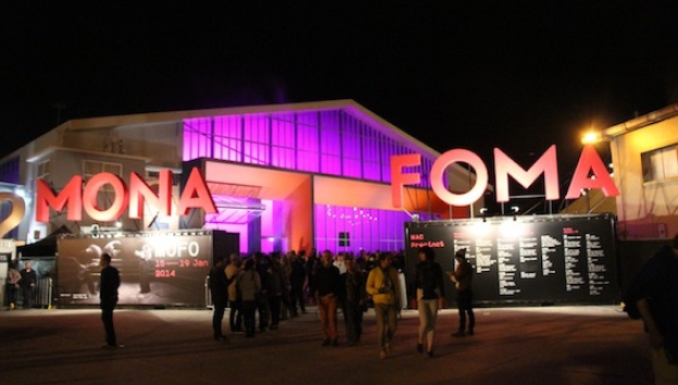 Exterior view of Mona Foma entrance at night as people walk in with purple and red lights illuminating the front signage