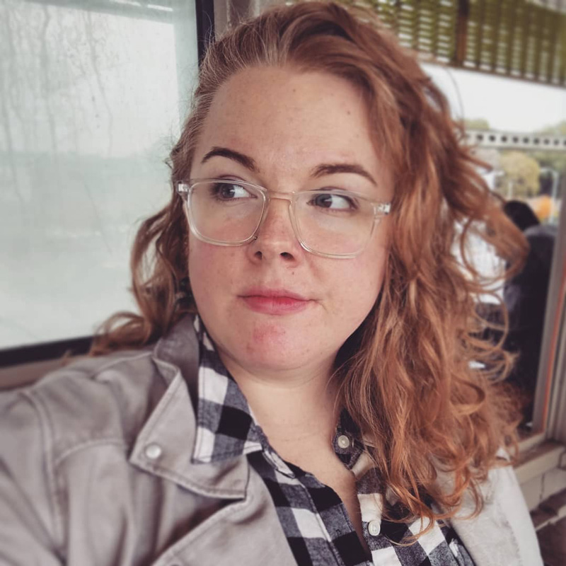 A fat woman wearing clear-framed glasses, a black and white checkered shirt, and wavy red hair.