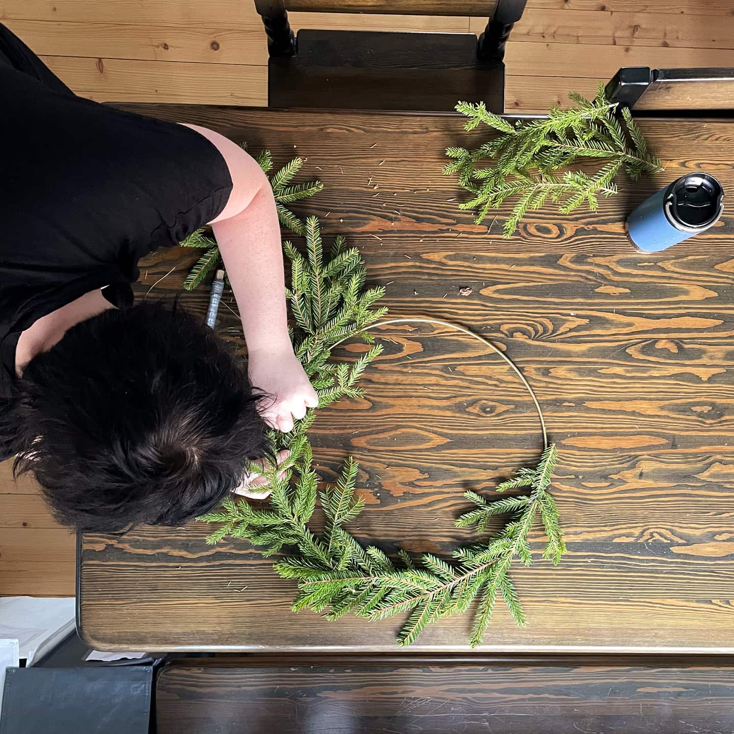Top-down view over a  kitchen table. A woman is tieing a wreath using spruce twigs.