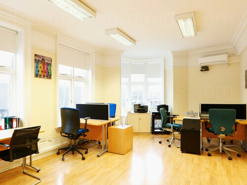 Well lit shared office workspace featuring wooden floors and desks with black office chairs under strip lights Microsoft Teams background