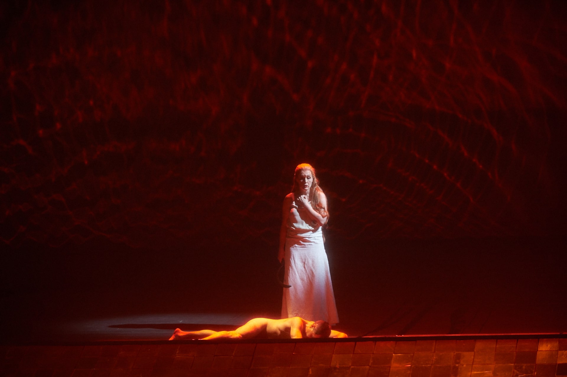 Woman in white clutches sickle over body of naked man, lit by shimmering red light overhead.