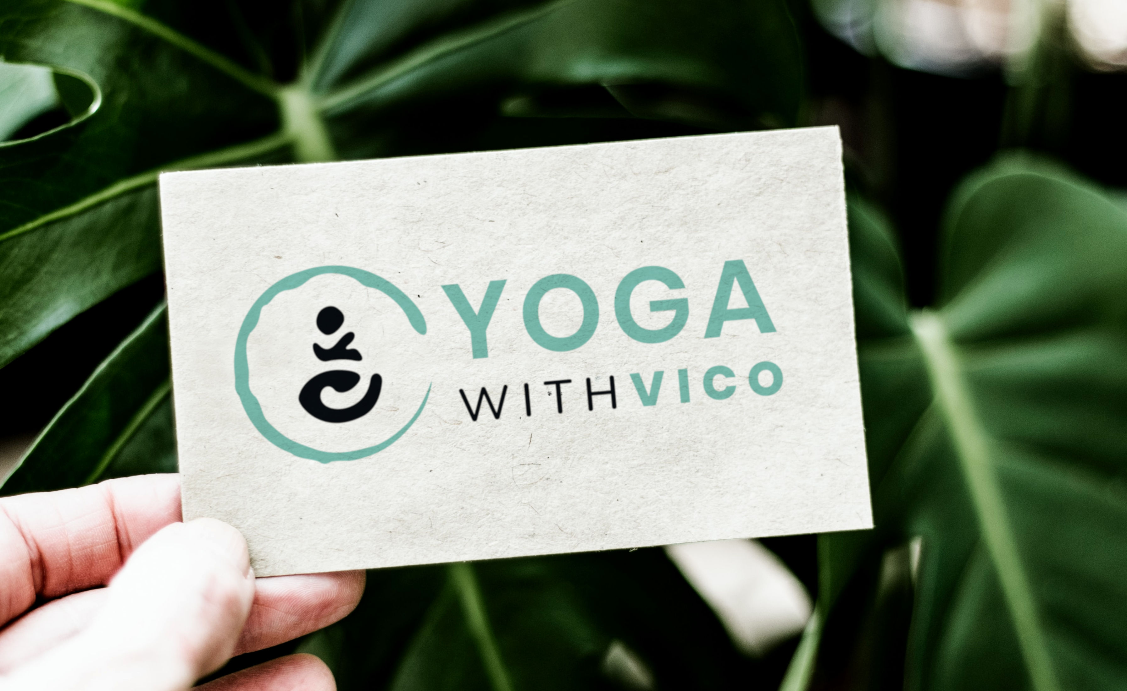 Yoga with Vico's rectangular logo design shown on a business card made from recycled paper. The card is held by a hand in front of green plant leaves.
