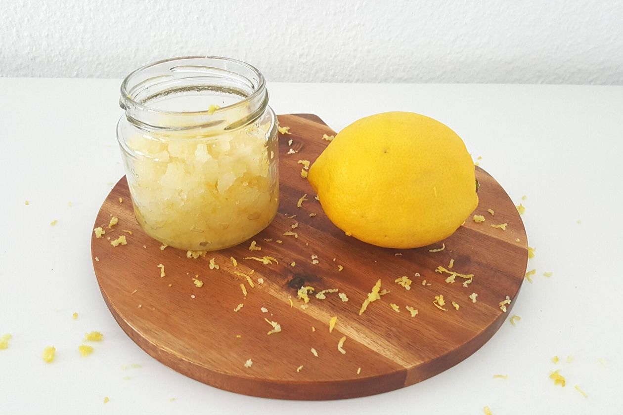 Das müllfreie Zitronen-Zucker Peeling in einem kleinen Schraubglas. Dieses ist auf einem Brett plaziert, daneben liegt eine Zitrone, drum herum ist Zitronenschale verstreut.