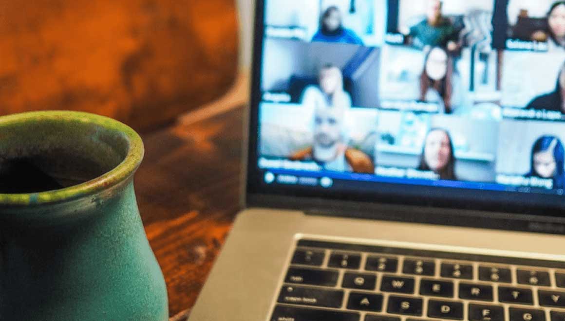 View of an online meeting on a laptop screen next to a mug of tea on a dining room table.