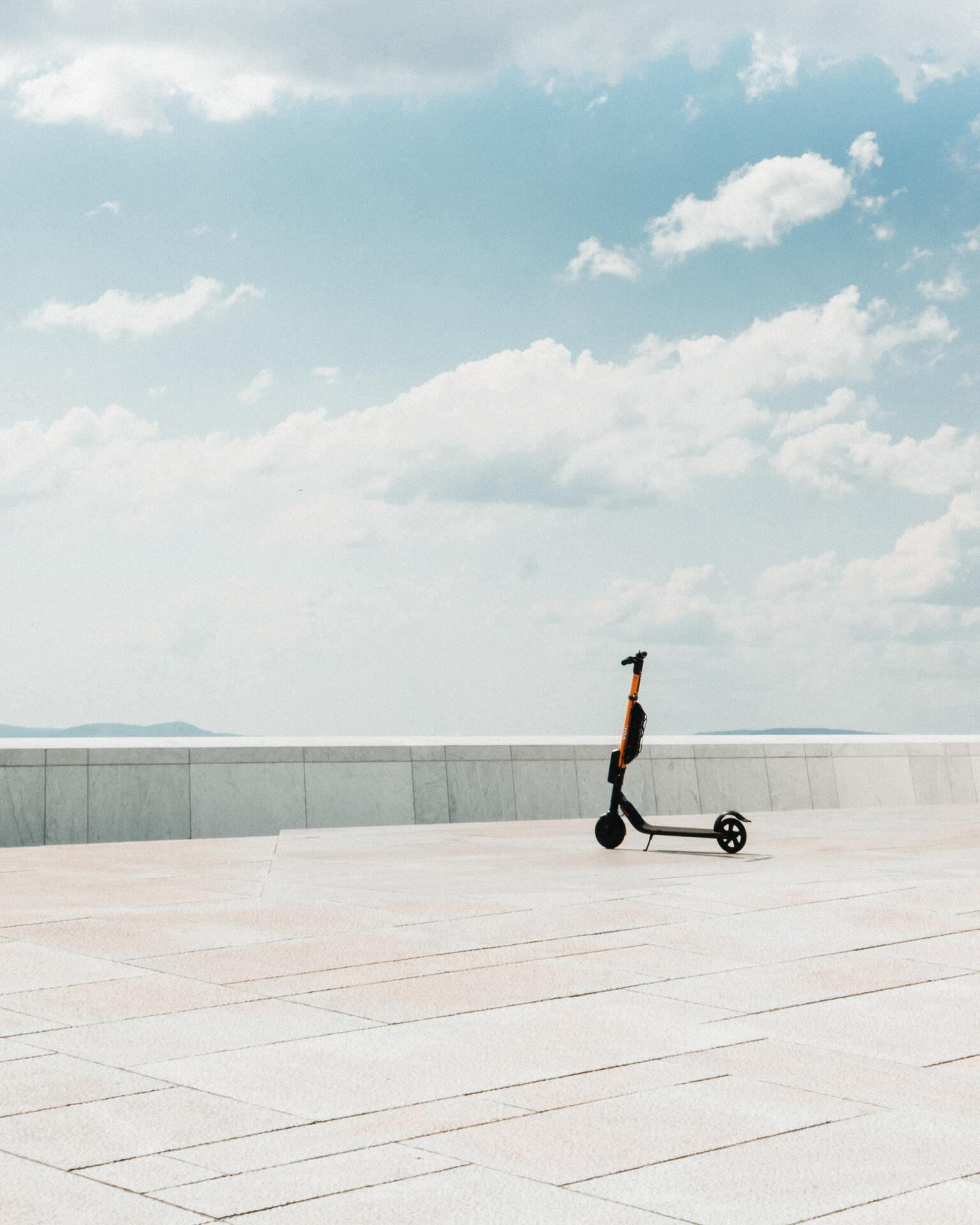 A red and black kick-scooter standing on top of an empty rooftop with an open and cloudy sky.