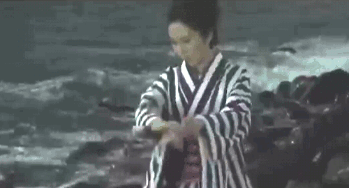 An animated gif of a scene from the movie 'Lady Snowblood' of a woman in a kimono killing a man with a katana along a rocky shore with waves crashing in.