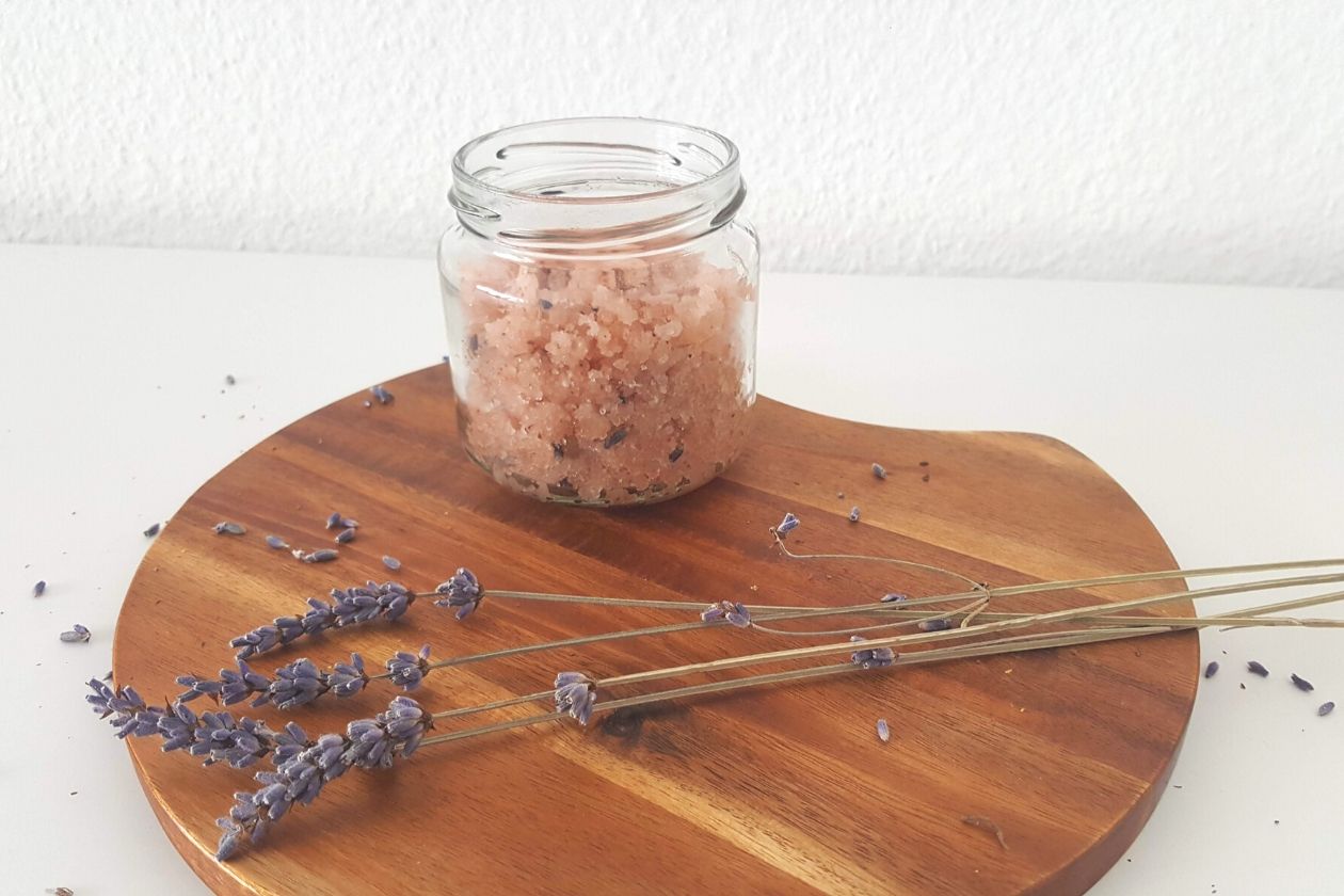 Das nachhaltige Lavendel-Salz Peeling in einem kleinen Schraubglas. Es ist auf einem Brett plaziert, davor liegen ein paar Lavendelzweige, drum herum sind Lavendelblüten verstreut.