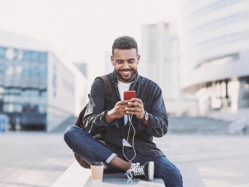 A smiling person sits on a wall in an urban setting looking at a smartphone screen.