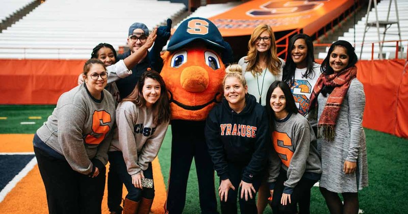 Syracuse students posing for a photo with their mascot, Otto the Orange, at a school sports facility