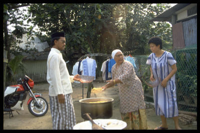 A Malay lady cooks in a big pot during Hari Raya Haji, in an open area within Kampong Sarhad. Two adults look on while they ready big empty bowls in their hands.