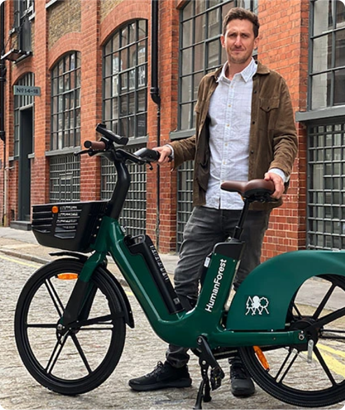 Human Forest card image with Agustin standing infront a brick building with a green human forest okai ebike.