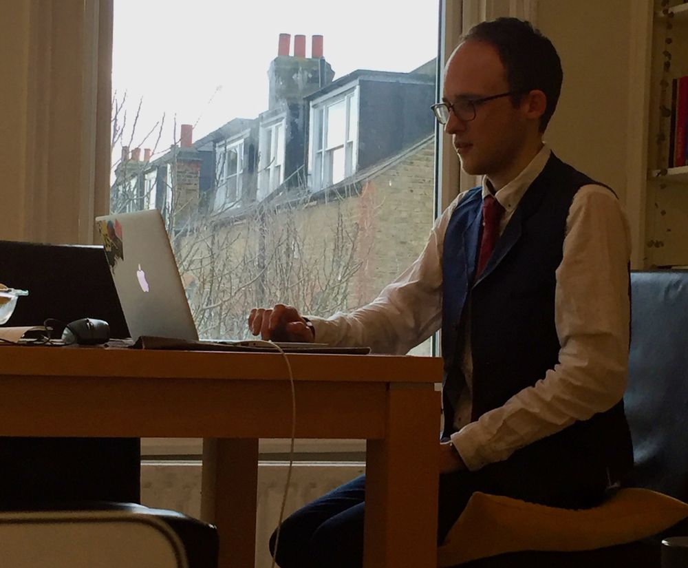 A photo of me sat at a dining table working. Taken in secret from the point of view of R, working from the sofa. I'm wearing a suit for fancy formal friday. I have no beard. I look weirdly young? I'm sat on a cushion because of the uncomfortable dining chair. Working from a 13 inch Macbook.