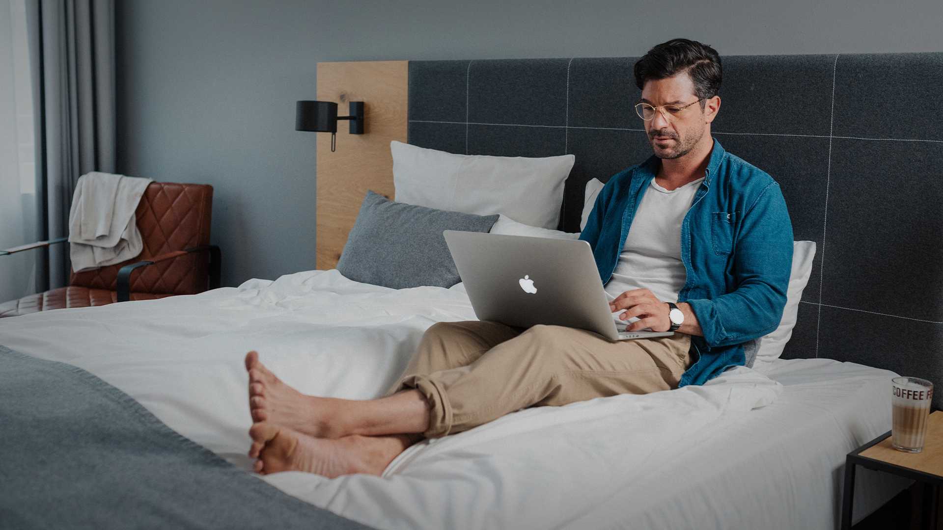 Mann sitzt auf seinem Hotelbett und arbeitet am Laptop