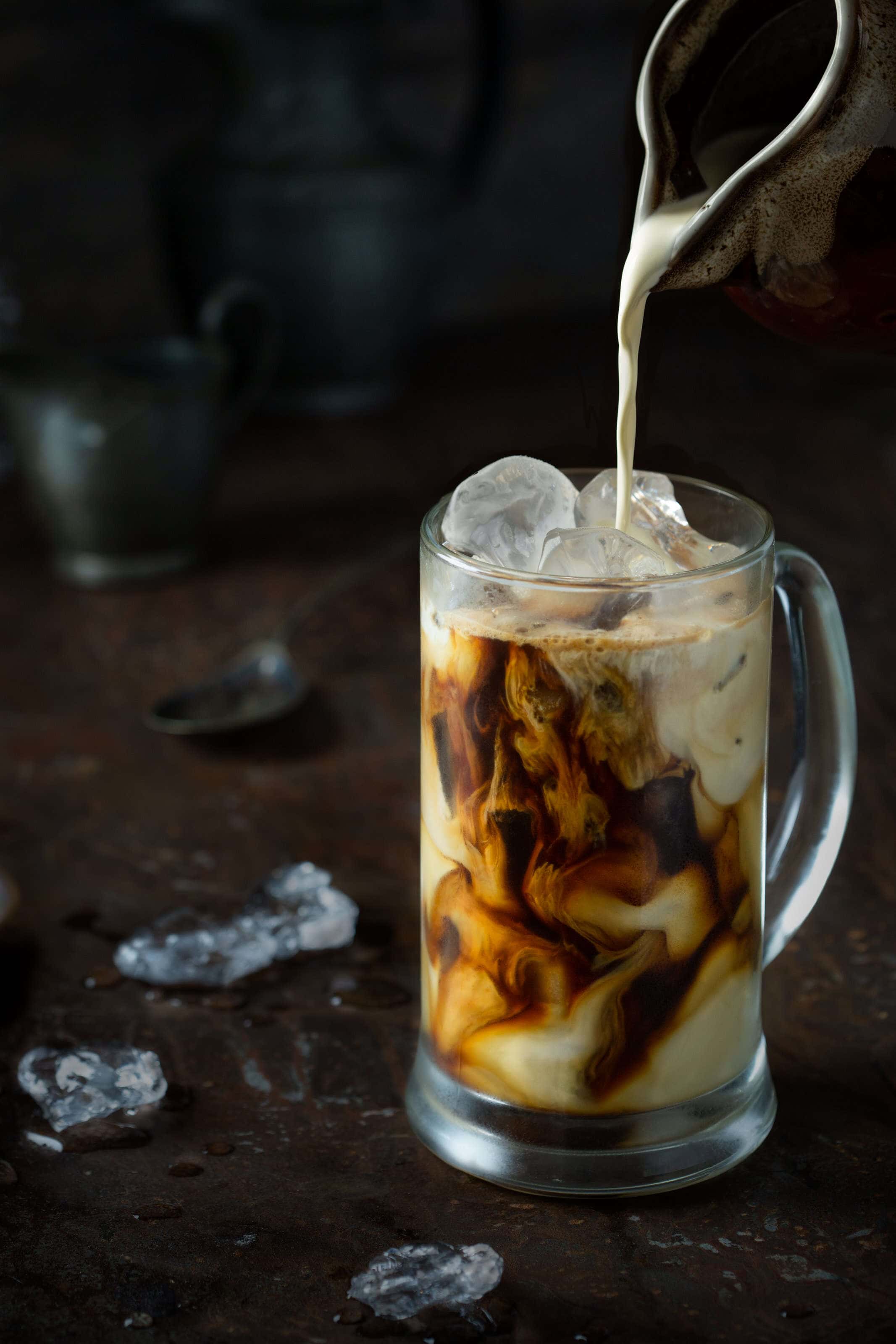 Iced coffee with cream being pored to create swirls through the coffee on a dark background.
