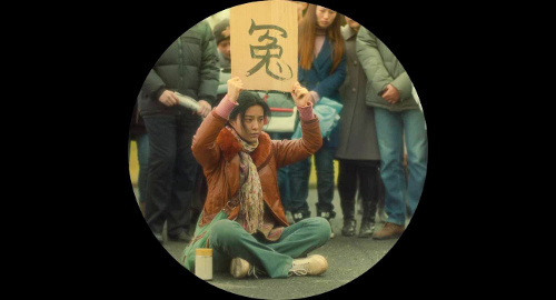 A screenshot from the film 'I Am Not Pan Jinlian' with a circular viewport showing a woman sitting on the ground cross-legged with a carboard sign above her head with the Chinese character written for 'Injustice'.