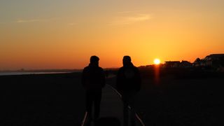 Two men walking on a board walk with orange skies behind then as the sun sets behind rooftops.