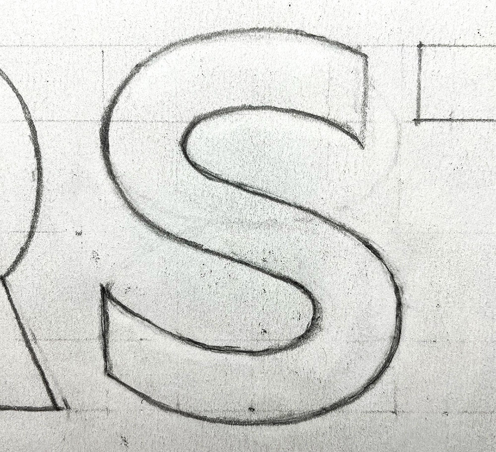 Hand drawn sans serif letter s in pencil on paper. The lines have been erased and redrawn many times so there’s a lot of smudges. The paper is white but it’s almost a light gray from all the erasing. Parts of other letters can be seen on the edges of the photo. 