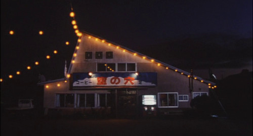 A screenshot showing a small coffeeshop at night with holiday lights strung up in front. The name of the store reads 'Hole in the Sky' in Japanese. From the movie 'Hole in the Sky'.