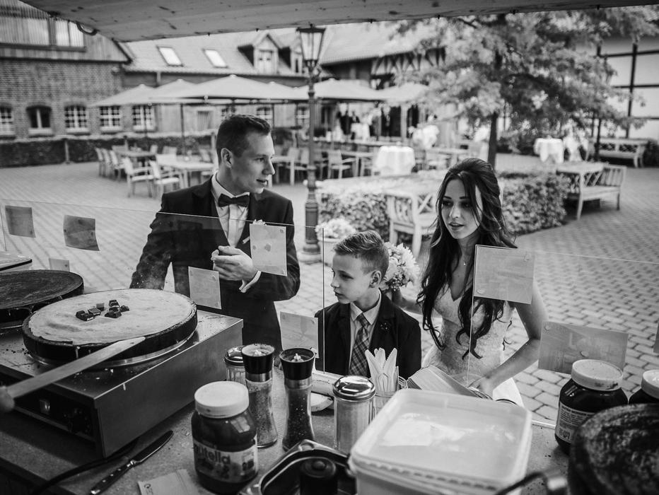 Brautpaar mit einem Kind schaut voller Vorfreude bei der Zubereitung eines Crêpe mit Kinderriegel zu