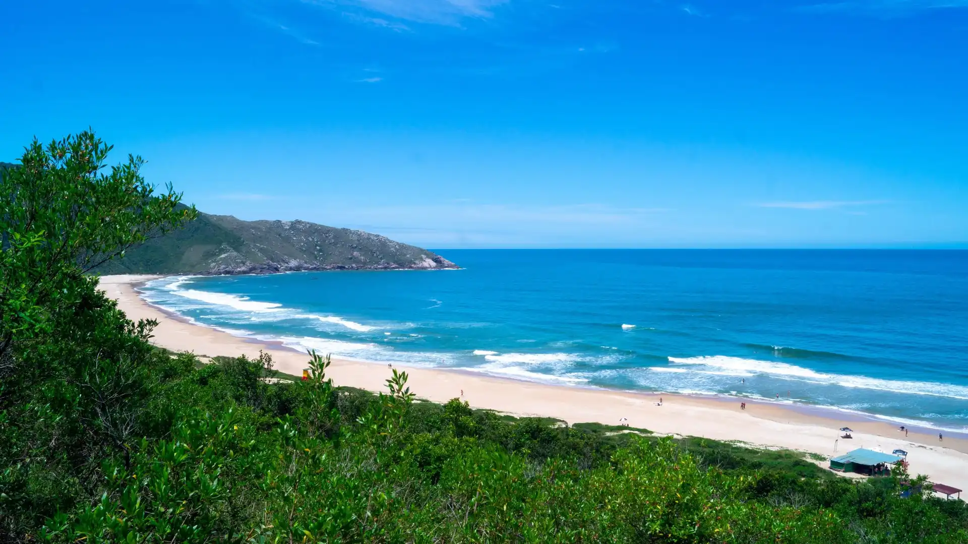 Vista da praia da Lagoinha do Leste, com par azul a direita e vegetação a esquerda. Ao fundo se vê o céu azul.