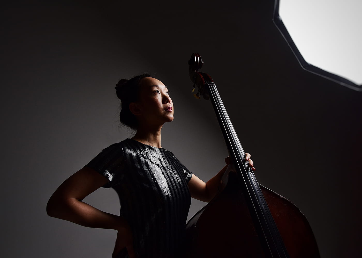 moody studio portrait from the side of Linda May Han Oh holding her double bass with a studio light over her head casting a harsh shadow