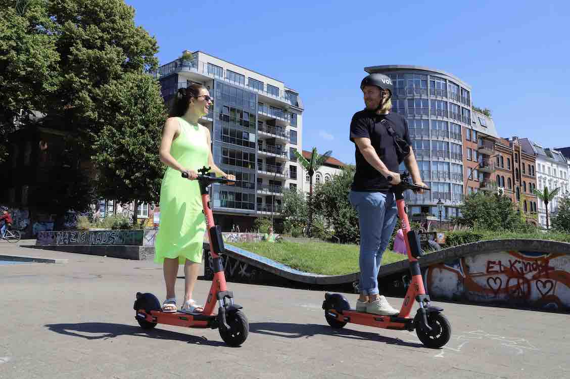 A male and female caucasian persons smiling and riding kickscooters in urban area.