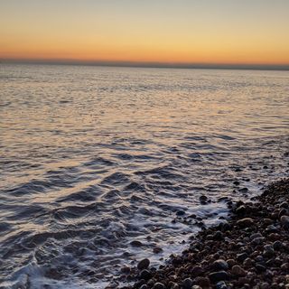 Calm sea with small wave breaking onto shingle and an orange sky.