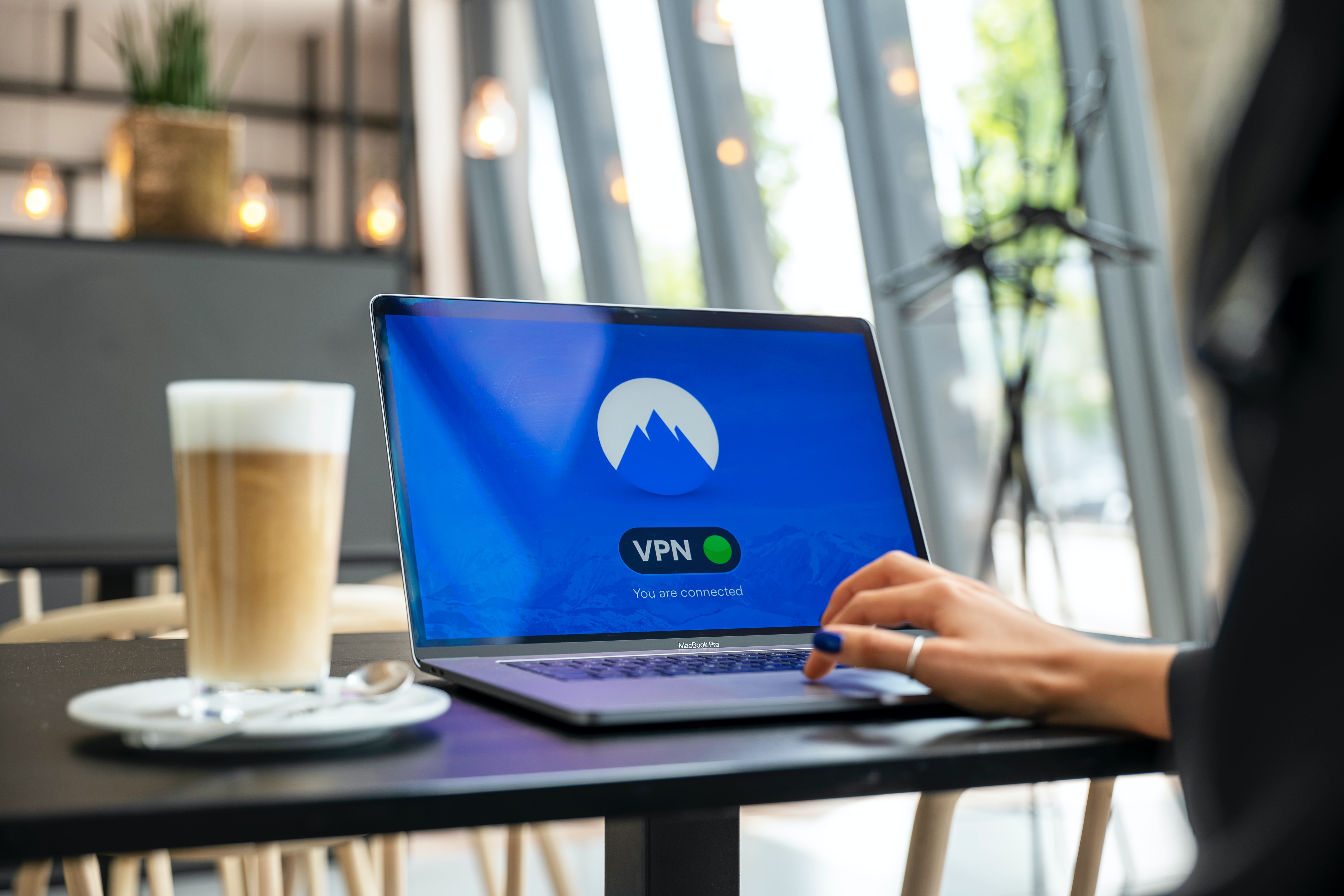 A woman sitting at a cafe counter with a latte and a laptop prominently showing a connected VPN on it.