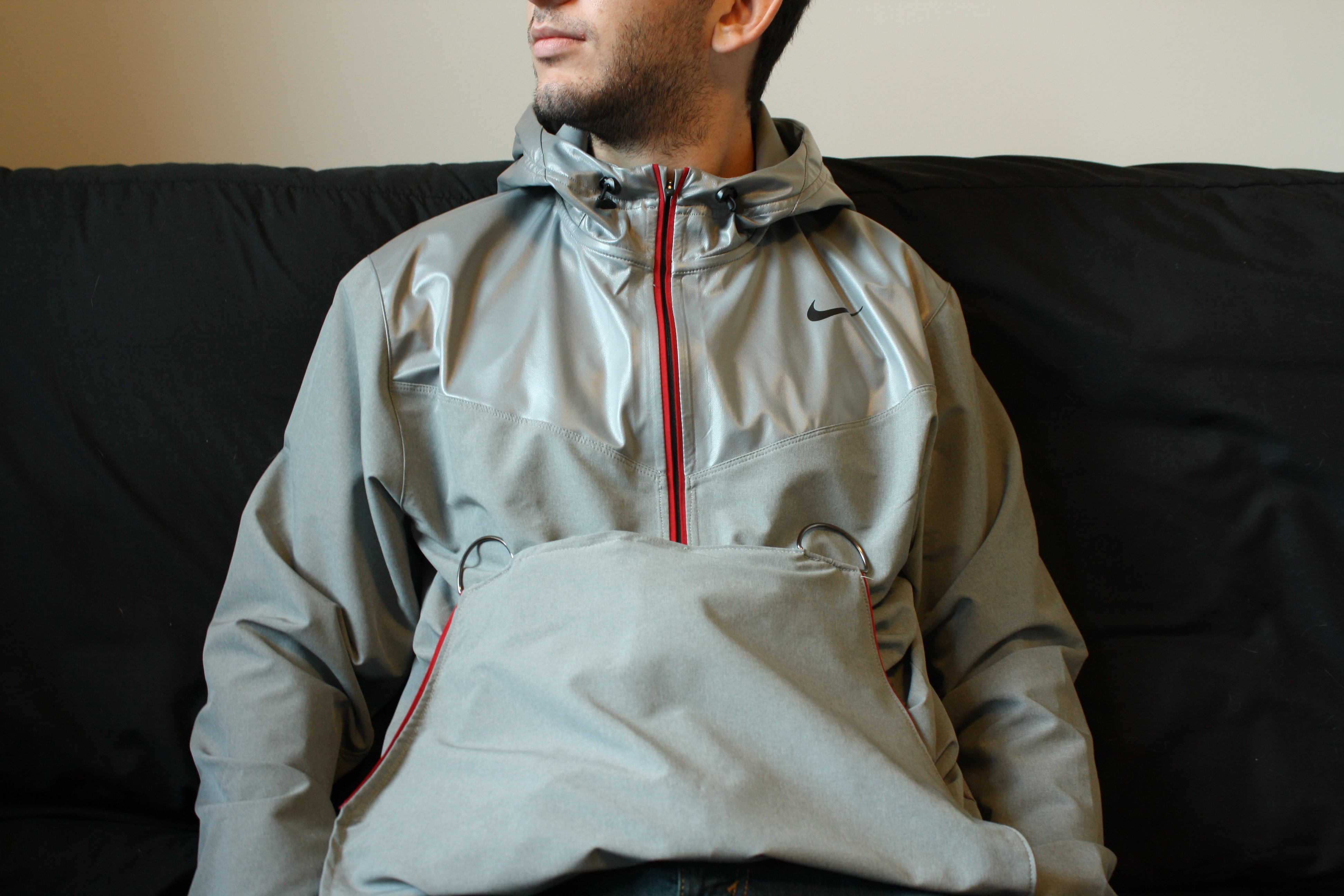 The RAYN jacket on a young man, seated. It's an altered Nike gray sports jacket with hoodie and zipper, but it's been radically customized in ways that are invisible at first glance.