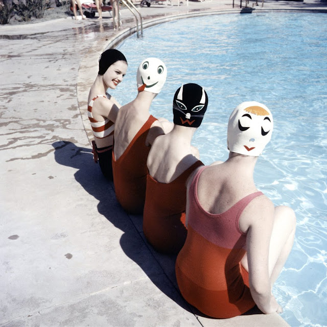 girls in bathing suits and vintage swimming caps printed with faces by a swimming pool