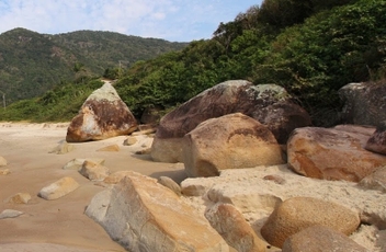 Foto da areia com grandes pedras em tons amarelados e morro ao fundo e pequena faixa de praia a esquerda