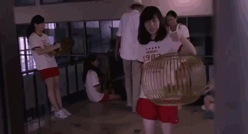 An animated gif from the movie 'Mary is Happy, Mary is Happy' of a girl in a red and white school uniform t-shirt walking towards the camera holding a round birdcage.