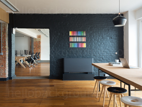 Contemporary wooden floored shared workspace containing wooden table and fabric topped stools with doorway view of desks Google Meet background