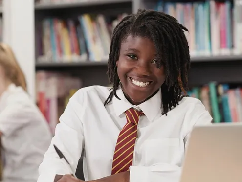 A student on a laptop in a classroom, smiling at the camera