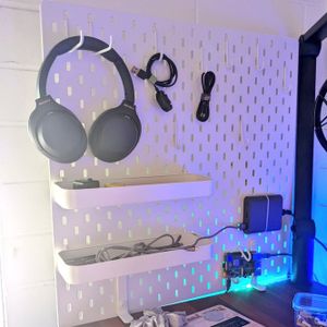 A white pegboard attached to the end of the desk against the wall, with several hooks and trays holding cables and a set of headphones