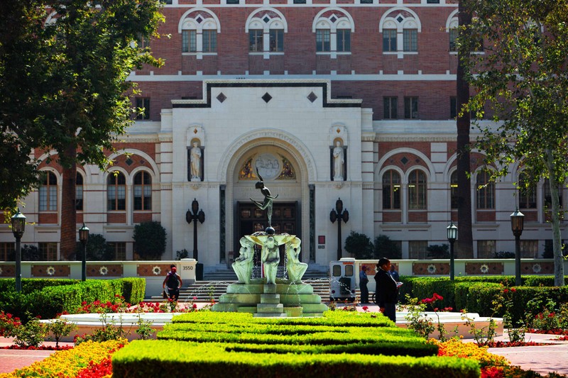 A USC campus building with a statue at the entrance and a fountain and gardens in the foreground