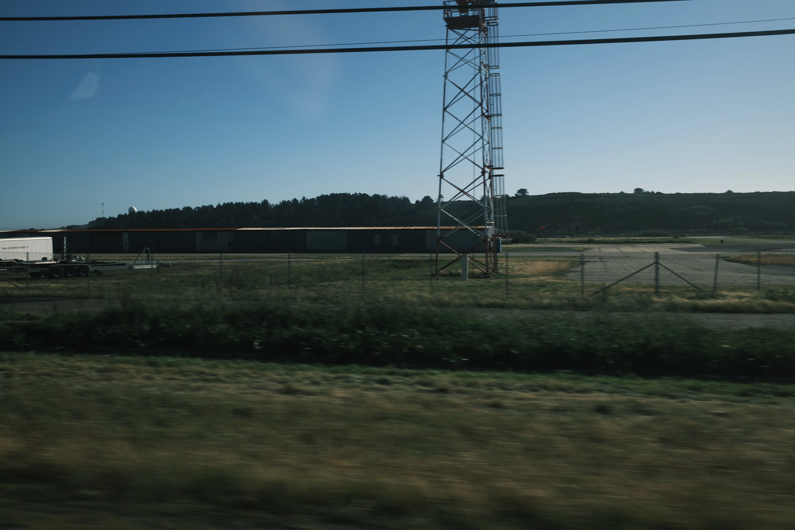 A blurry photo taken from a car of a field with power lines and a large, squat building in the background