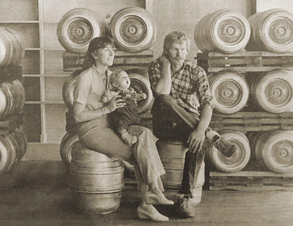 Sepia toned photo of Tom Baune and Beth Hartwell sitting on kegs with kegs stacked up in the background. Beth is holding a baby.