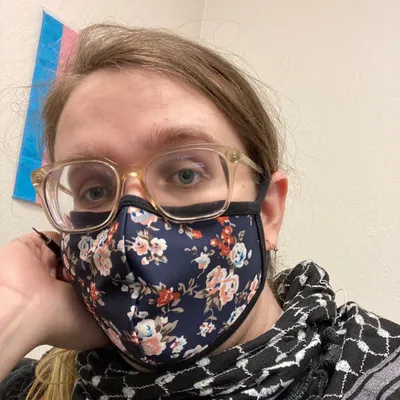 A portrait of Sarah Robin wearing a floral facemask and thick eyeglasses with her brown hair tied back in a ponytail, taken in front of a transgender pride flag