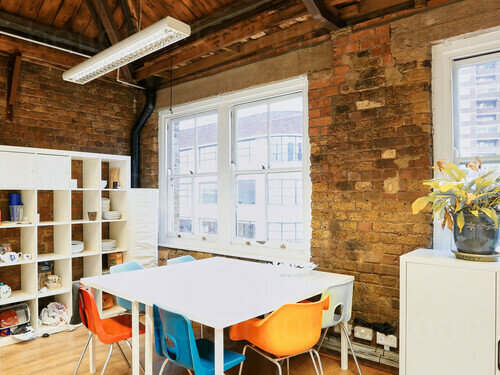 Intimate meeting room with bare brick walls containing square white table with multi-coloured chairs next to white bookcase Skype background