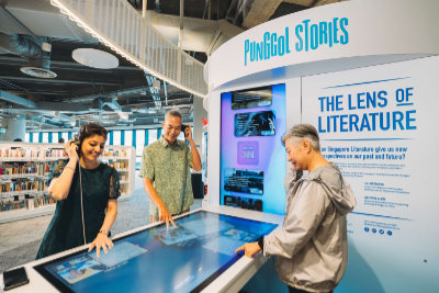 A photo of the Lens of Literature exhibition. It features an interactive multimedia table with an adjacent screen. Visitors are exploring the content displayed on the multimedia table while holding its sound handsets to their ears.