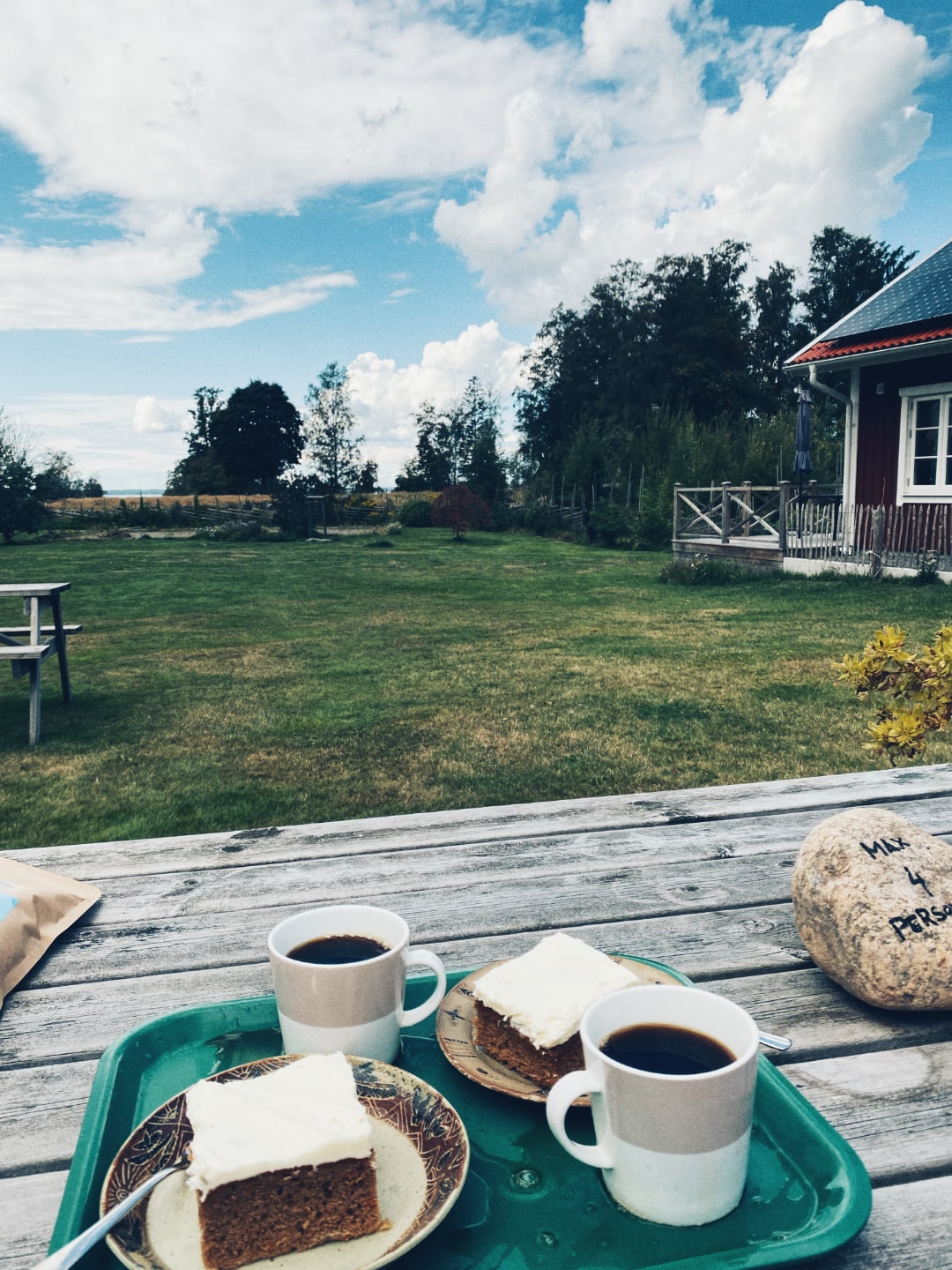Coffe and carrot cake on a wooden table outside in, what looks like, a garden. In the background, one can catch a glimpse of a roundpole fence.