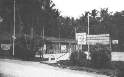A black and white photograph of the single-storey Kampong Sungei Tengah Community Centre and its outdoor basketball court amidst a coconut plantation.