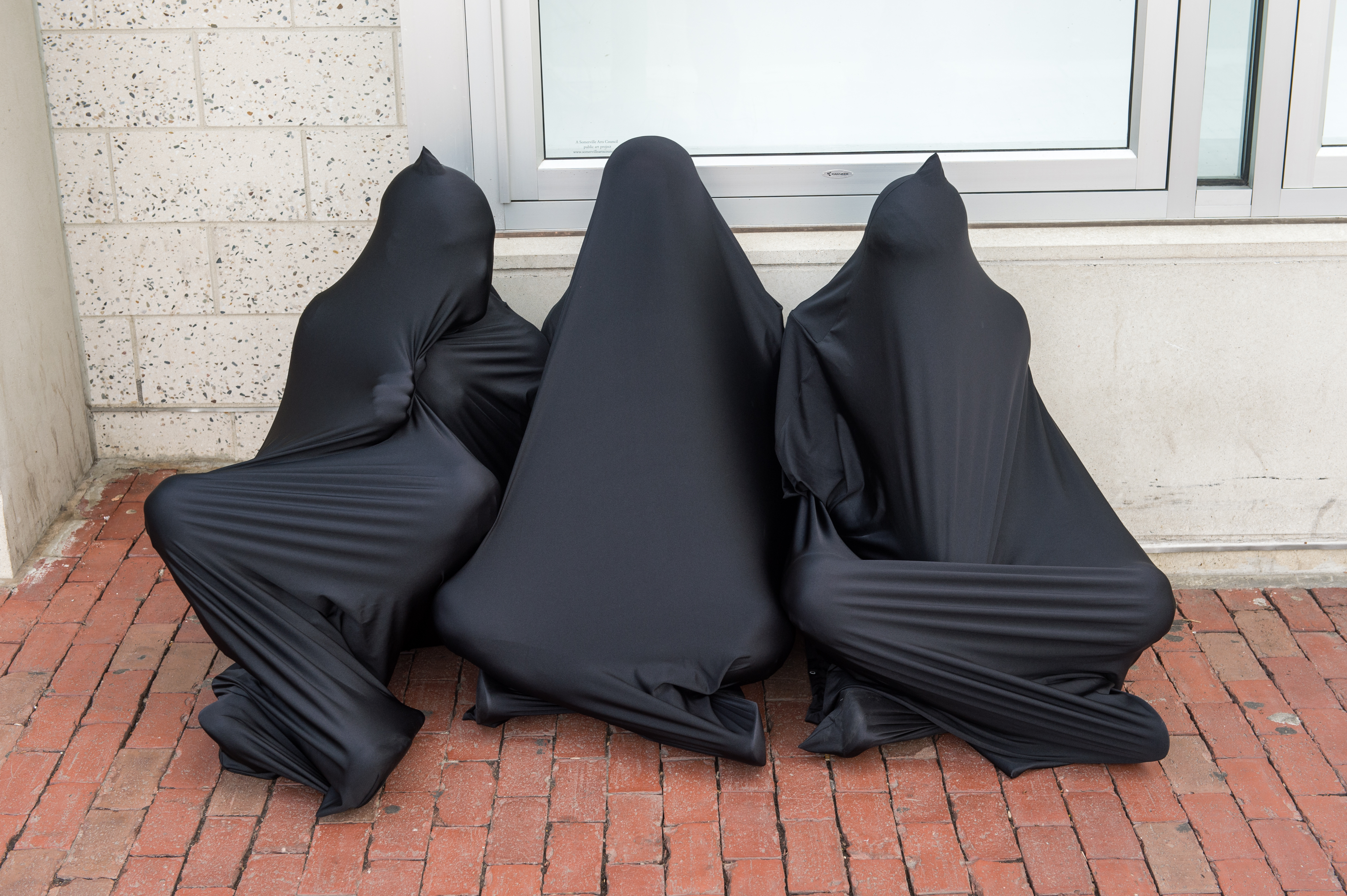 Three figures, seated outside, huddled together in separate black lycra body socks, enveloping their entire bodies.