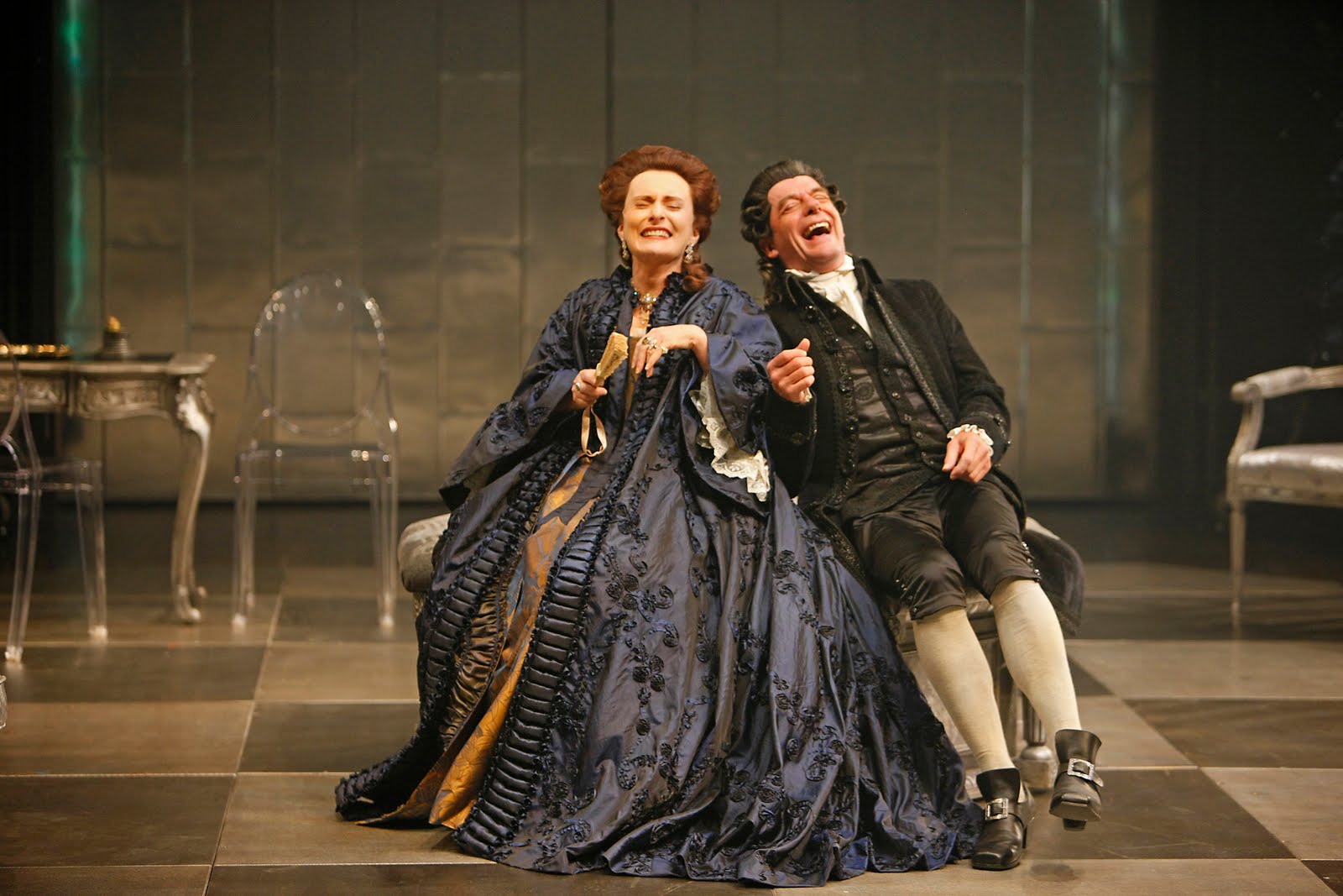 Woman in navy gown sits laughing with black-suited man on checkered floor.