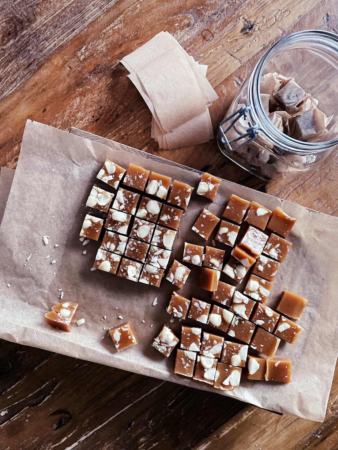 Chopped up pieces of caramel candy, laying on baking paper. There's also a glass jar with wrapped candies and wrapping paper.