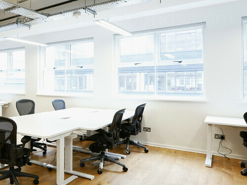 Simple and light shared office workspace containing white tables with black office chairs next to large windows Google Meet background