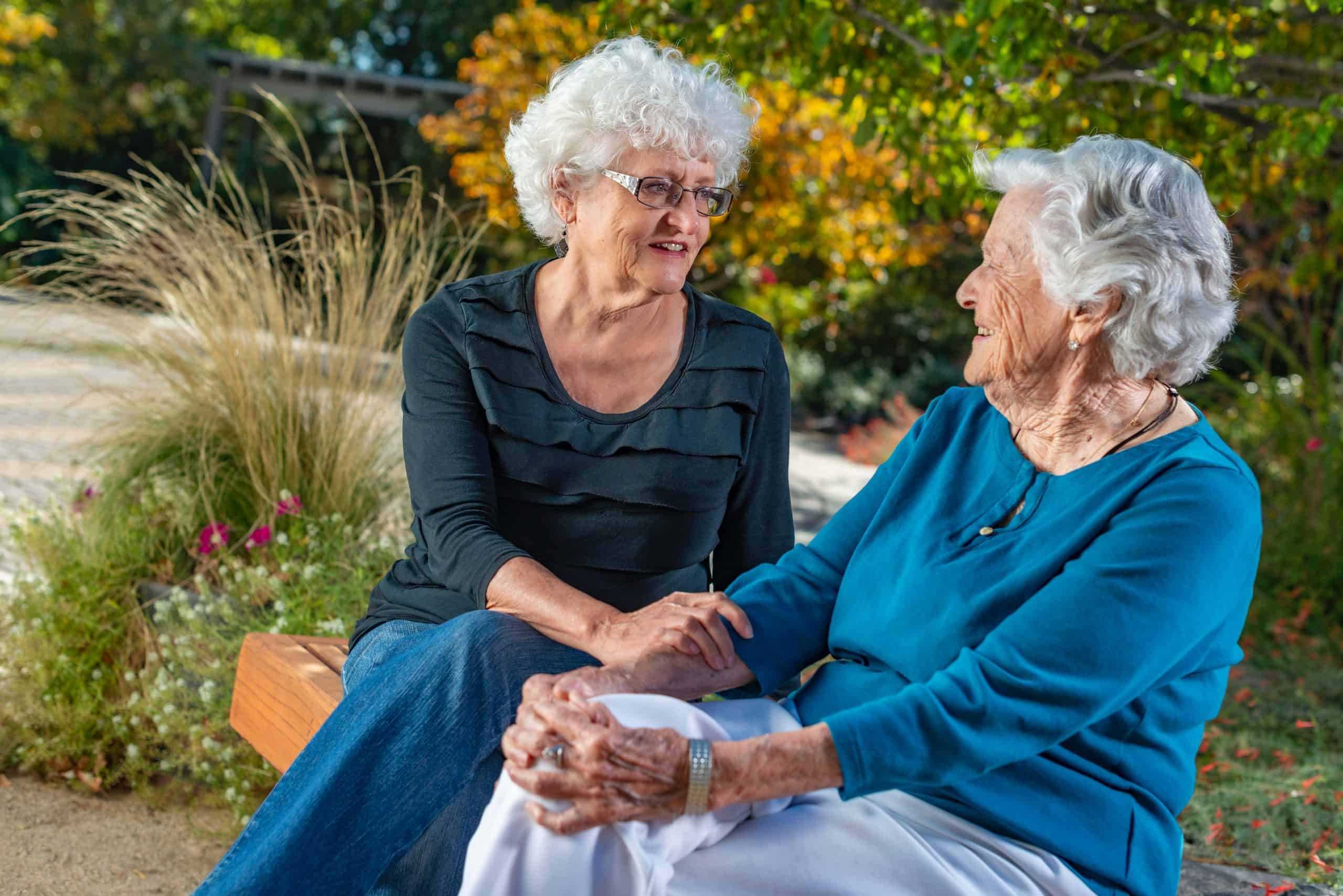Two elderly women talking and smiling on a bench in a garden